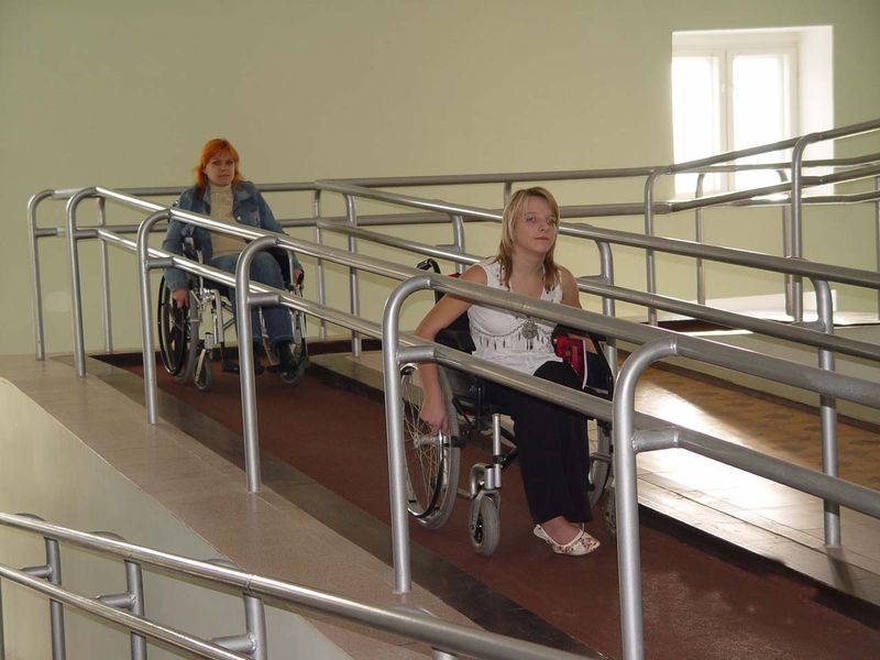 Цветное фото. На фото длинный пандус с перилами внутри помещения. По пандусу спускаются две молодые девушки на инвалидных колясках.